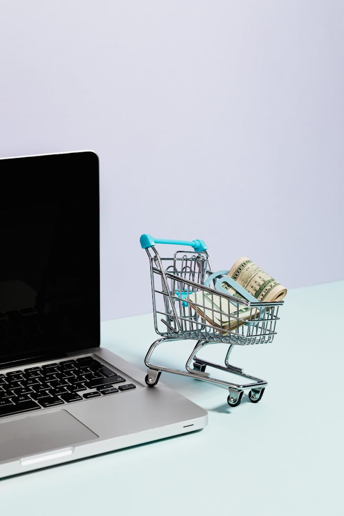 digital Mini shopping cart with rolled cash next to a laptop, representing online shopping.